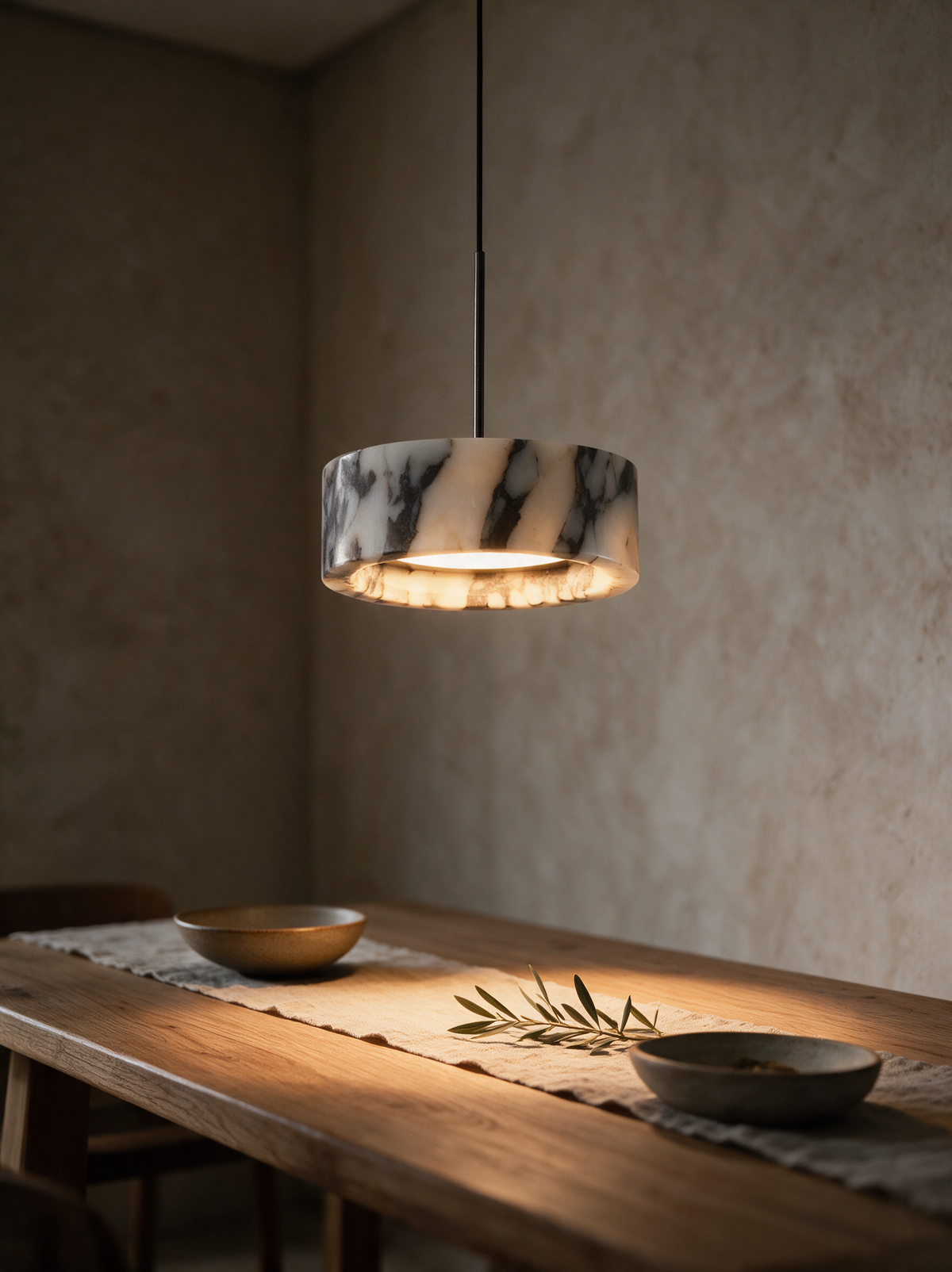 Aria Pendant Lamp in marble, switched on, hanging low over an oak dining table at dusk