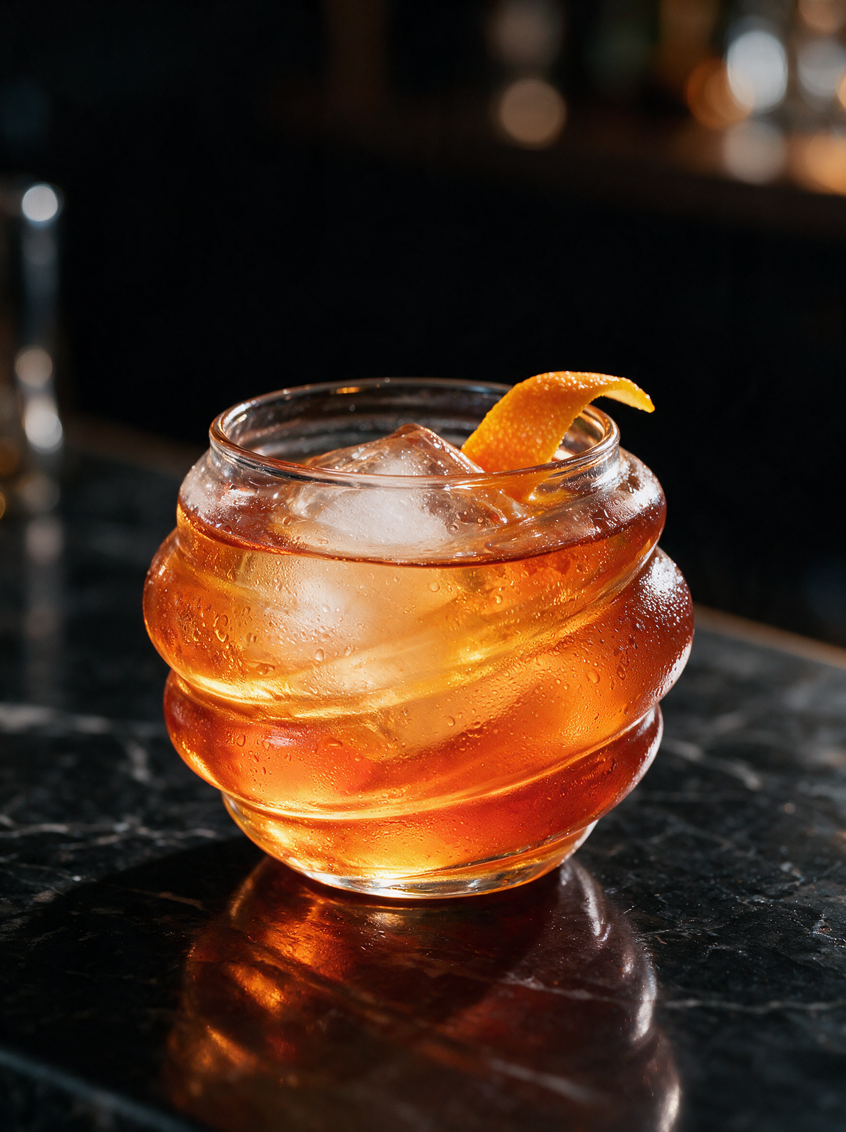 Onda Glass Cup holding an amber cocktail on a dark marble bar
